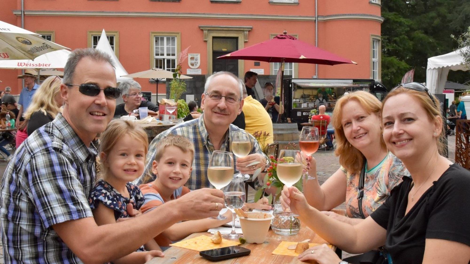 Eine Familie prostet dem Fotografen zu. Es sind vier Erwachsene und zwei Kinder. Im Hintergrund steht ein altes rotes Gebäude.