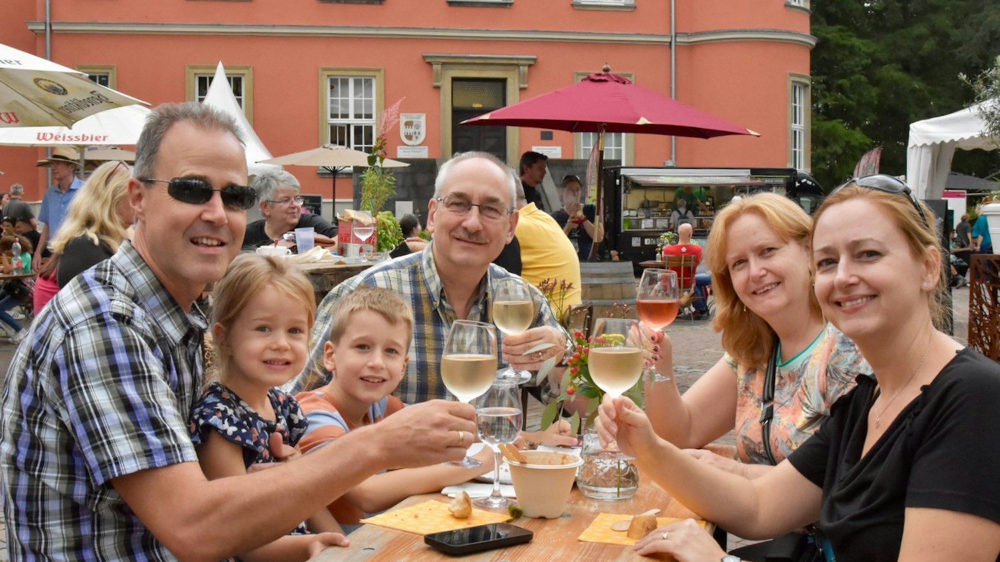 Eine Familie prostet dem Fotografen zu. Es sind vier Erwachsene und zwei Kinder. Im Hintergrund steht ein altes rotes Gebäude.