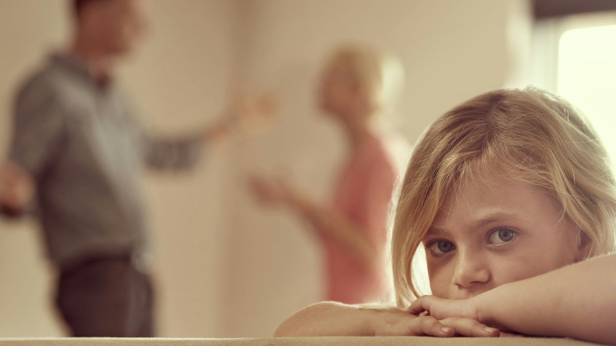 Ein blondes Mädchen schaut traurig in die Kamera, im Hintergrund streiten sich die Eltern.