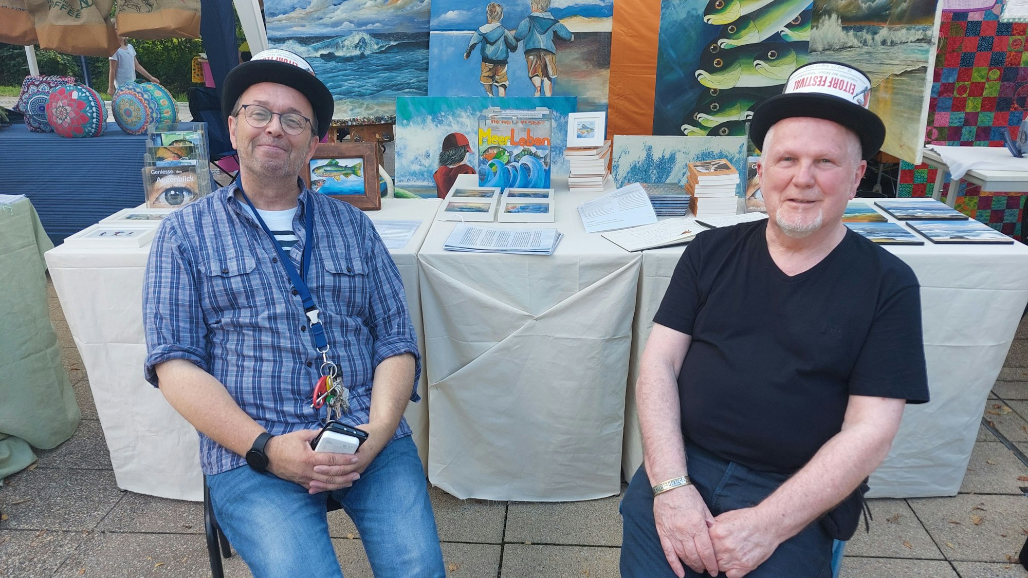 Zwei Männer mit Zylinder sitzen vor einem Tisch, auf dem Bilder zum Kauf angeboten werden.