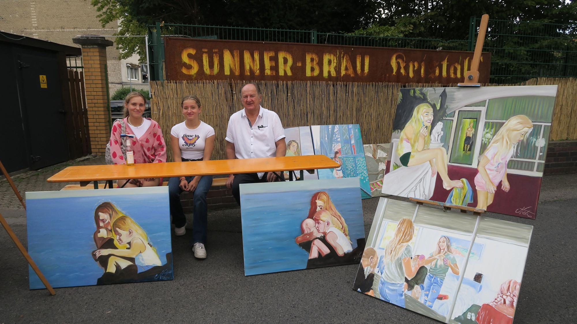 Drei Menschen sitzen an einem Stand mit Gemälden.