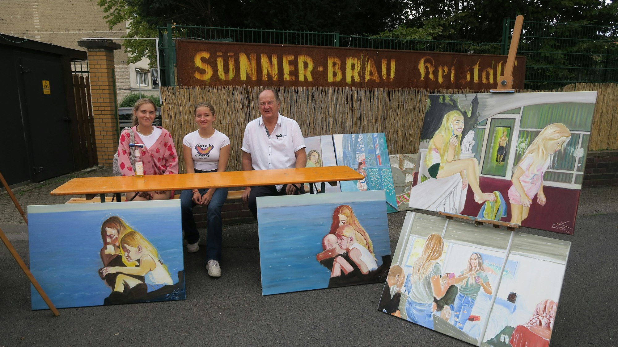 Drei Menschen sitzen an einem Stand mit Gemälden.