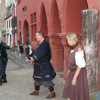 An einen Baumstamm gefesselt steht die überführte Händlerin Erika Rimkus. Ratsherr Sebastian Pöschke und Büttel Sven Kreitmann stehen daneben. Sie alle tragen mittelalterliche Kleidung. Eine Gruppe von Menschen in moderner Straßenkleidung steht davor, schaut und lacht. Mit dem Smartphone machen einige von ihnen Fotos.