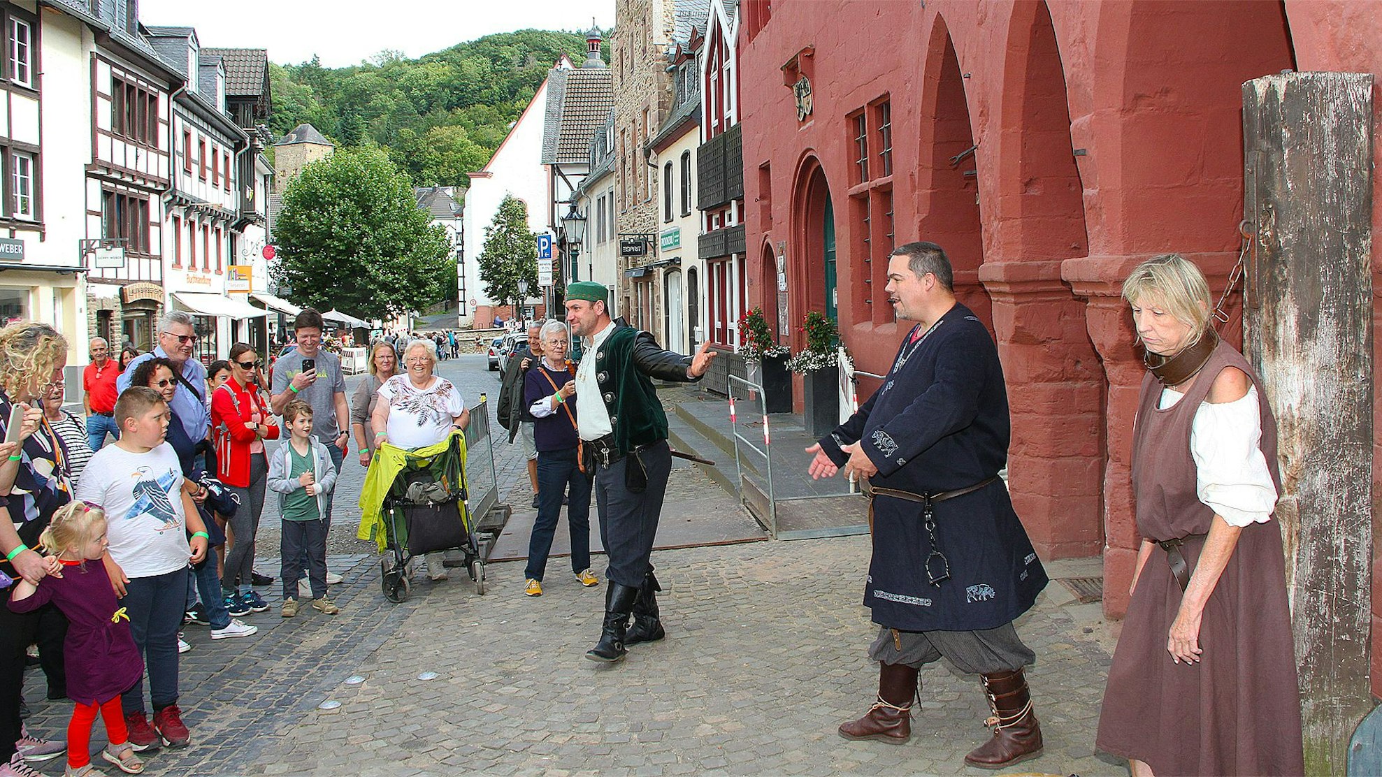 An einen Baumstamm gefesselt steht die überführte Händlerin Erika Rimkus. Ratsherr Sebastian Pöschke und Büttel Sven Kreitmann stehen daneben. Sie alle tragen mittelalterliche Kleidung. Eine Gruppe von Menschen in moderner Straßenkleidung steht davor, schaut und lacht. Mit dem Smartphone machen einige von ihnen Fotos.