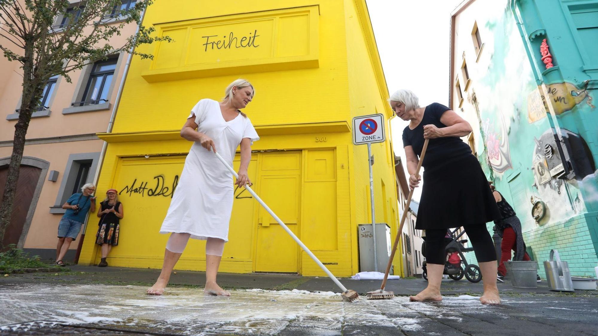 Zwei blonde Frauen schrubben den Asphalt mit Besen und Reinigungsmittel.