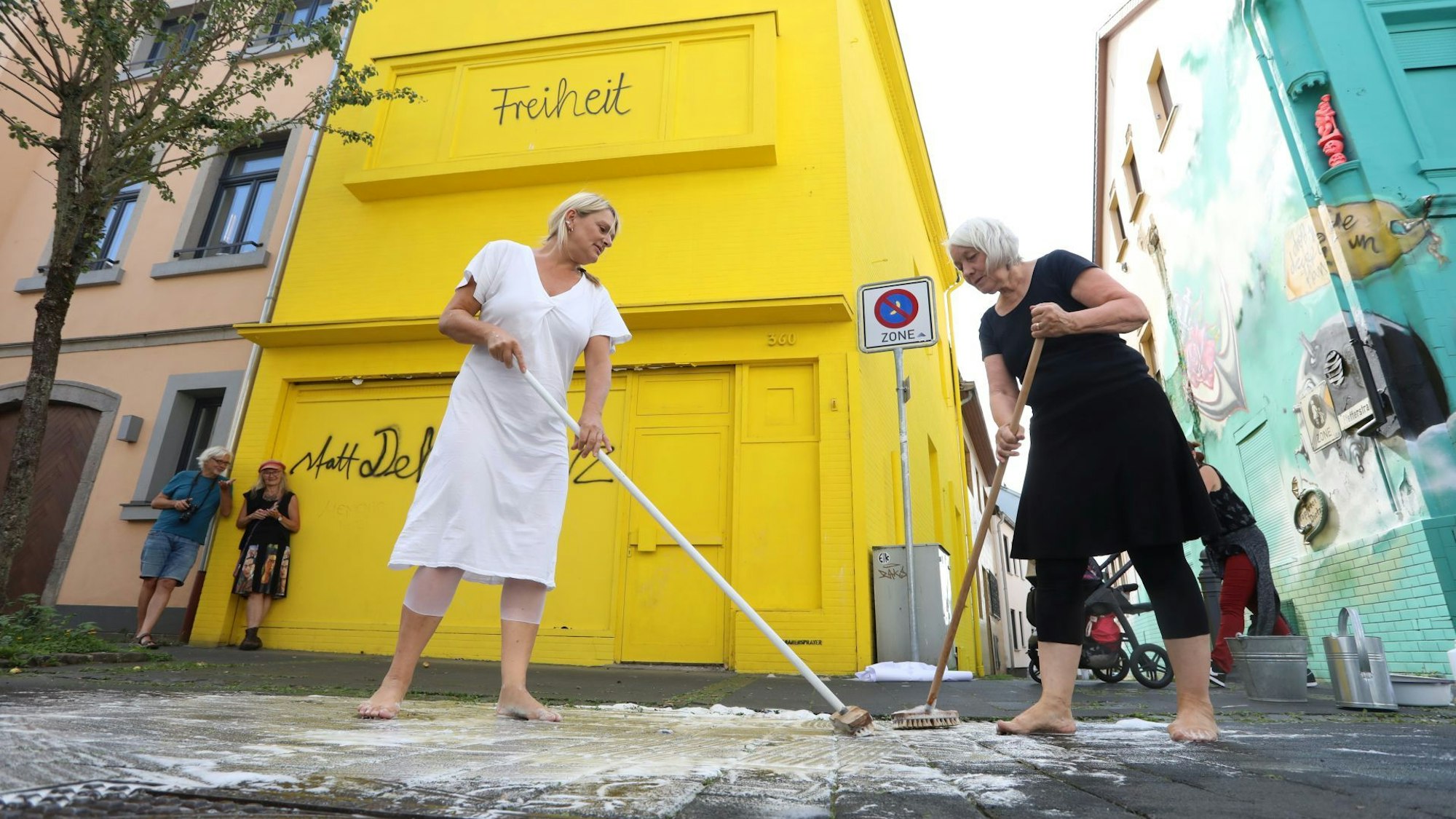 Zwei blonde Frauen schrubben den Asphalt mit Besen und Reinigungsmittel.