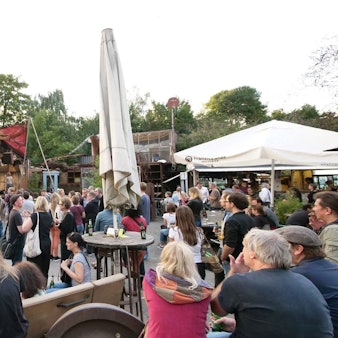 Auf einem Festival-Gelände mit Sonnenschirmen, Stehtischen und einer kleinen Bühne stehen viele Menschen.