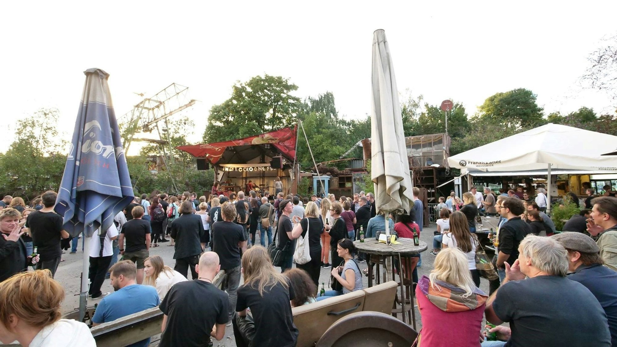 Auf einem Festival-Gelände mit Sonnenschirmen, Stehtischen und einer kleinen Bühne stehen viele Menschen.