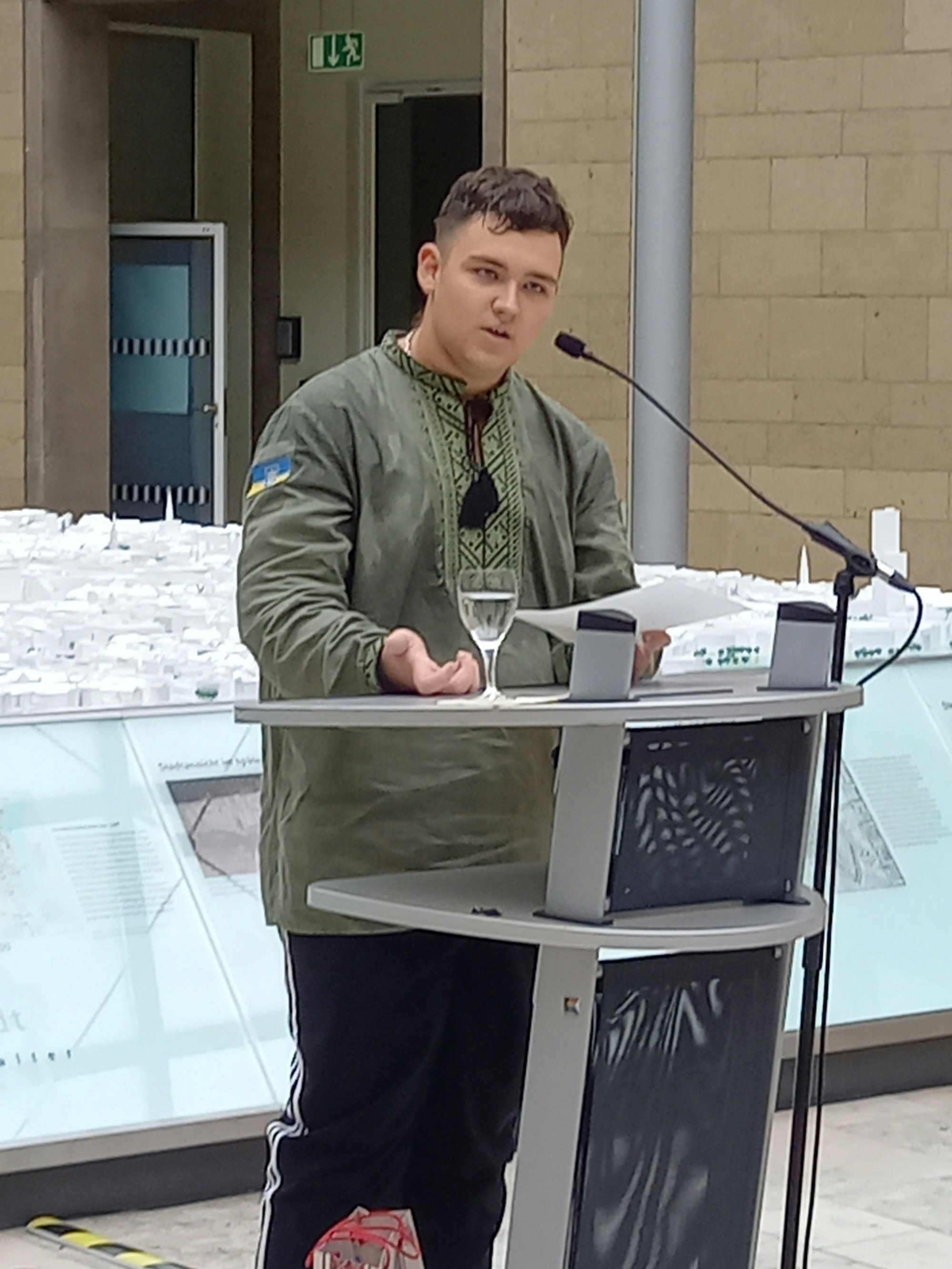 Ein junger Mann im dunkelgrünen Hemd mit der ukrainischen Fahne auf dem Oberarm steht an einem Rednerpult und spricht.