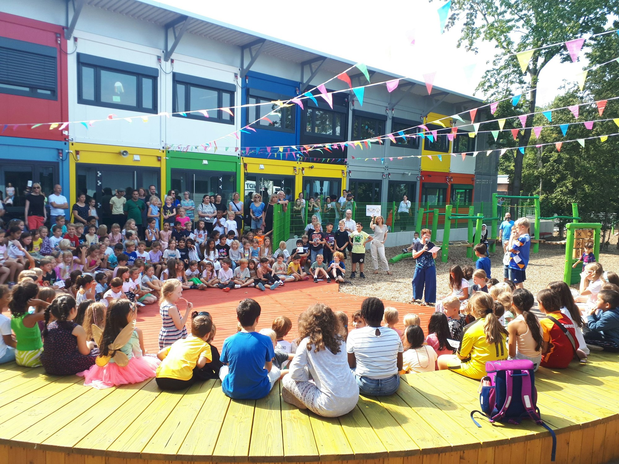 Viele Kinder sitzen im Halbkreis im Schulhof, eine Lehrerin spricht in ein Mikrofon.