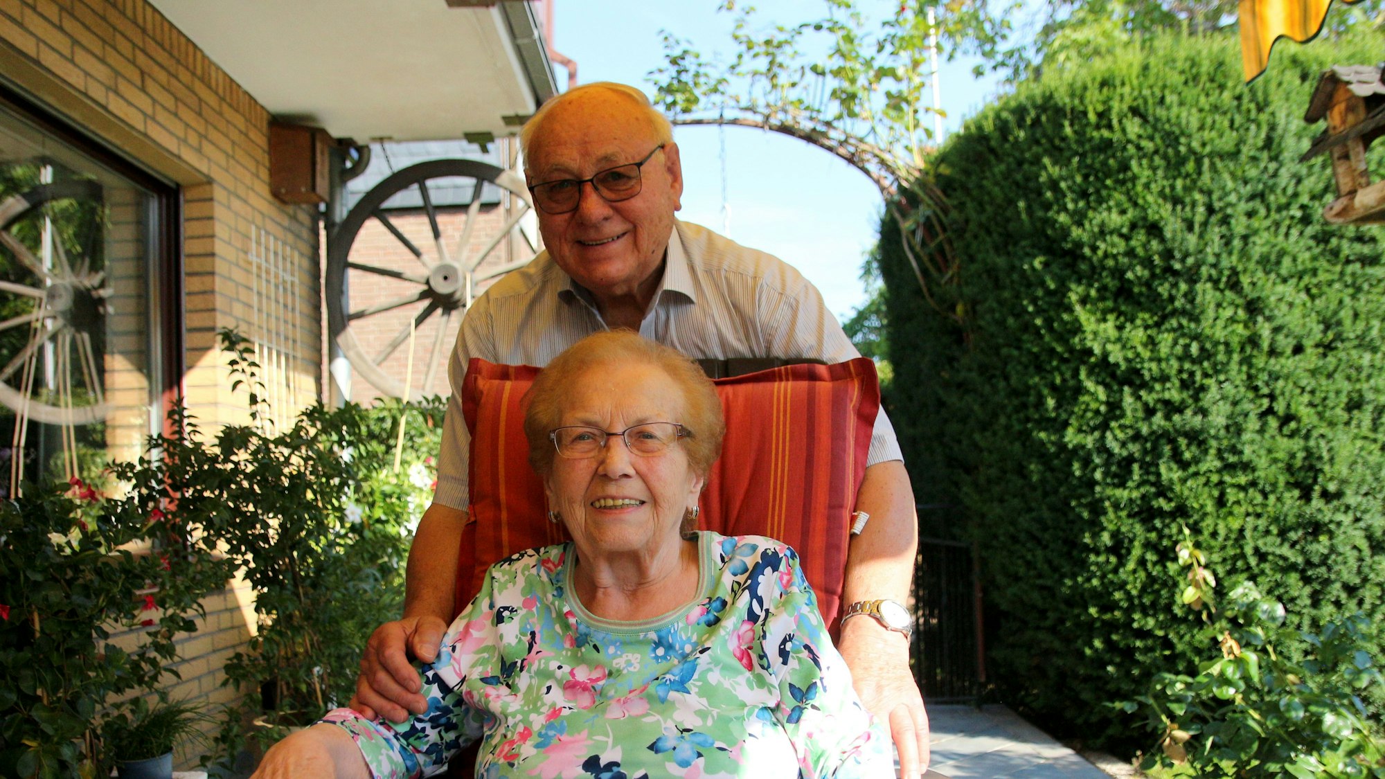 Dorothea Czwalinna sitzt in einem Gartenstuhl, ihr Mann Hans steht dahinter und legt seine Hände auf ihre Arme.