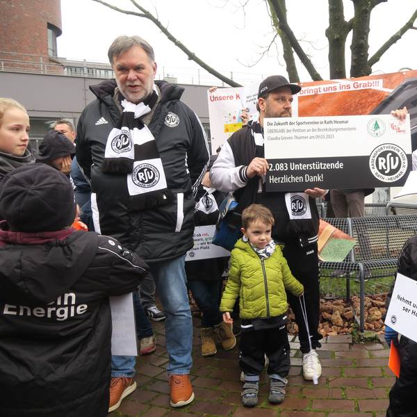 Männer und Kinder in Vereinsjacken und -schals übergeben einer Frau in weißer Jeansjacke einen groß aufgezogenen Petitions-Beleg.