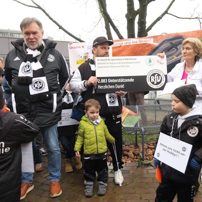 Männer und Kinder in Vereinsjacken und -schals übergeben einer Frau in weißer Jeansjacke einen groß aufgezogenen Petitions-Beleg.