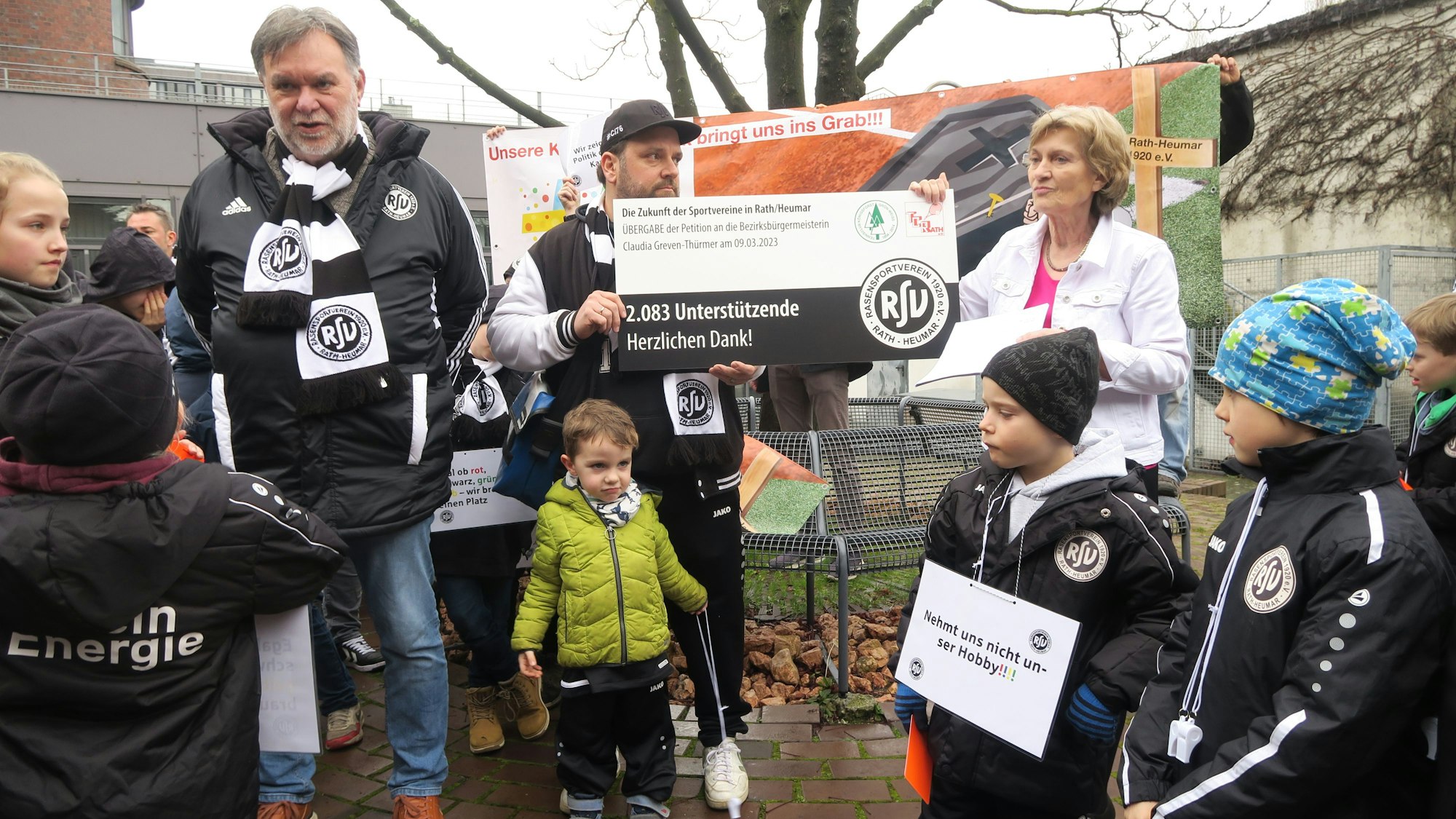 Männer und Kinder in Vereinsjacken und -schals übergeben einer Frau in weißer Jeansjacke einen groß aufgezogenen Petitions-Beleg.