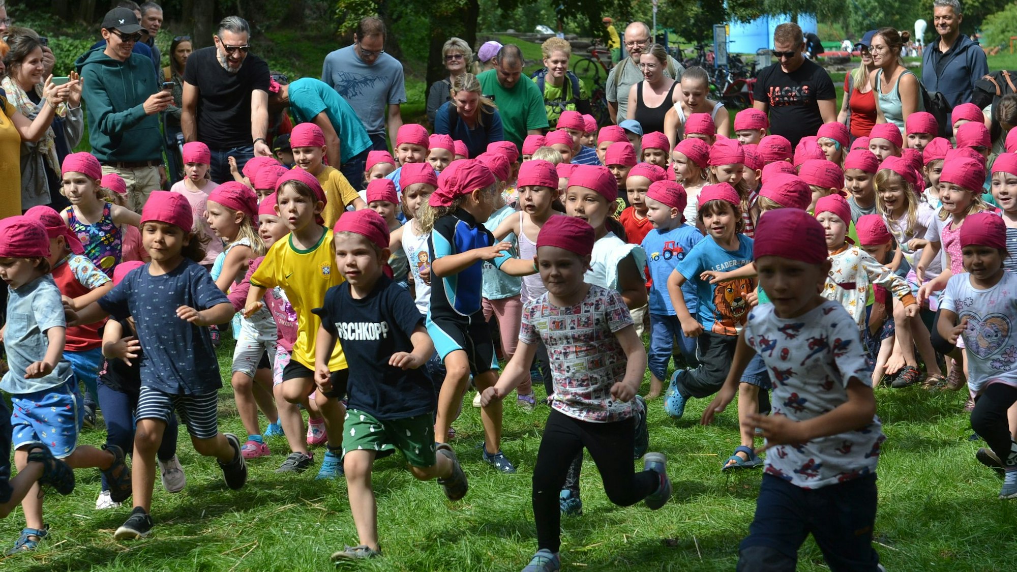 Eine große Gruppe Kinder läuft über eine Wiese. Sie alle tragen pinke Tücher um den Kopf. Im Hintergrund stehen einige Eltern.