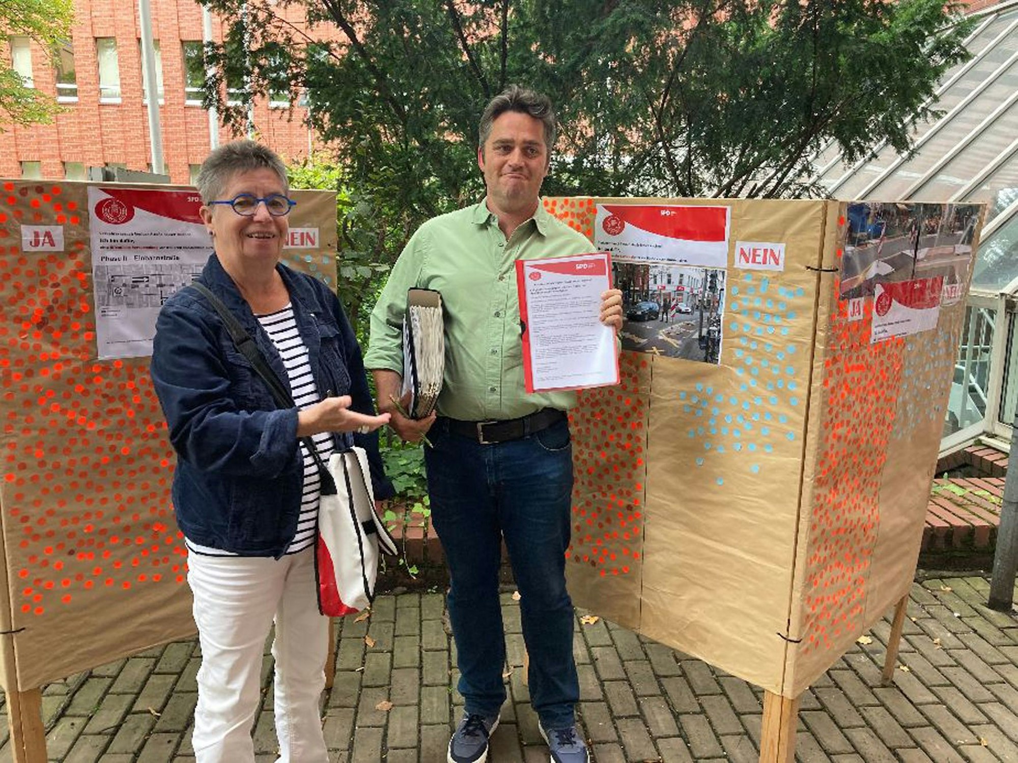 Eine Frau mit grauem kurzen Haar und Brille steht neben einem Mann in grünem Hemd und kurzem dunklen Haar. Der Mann hat Schnellhefter und Aktenordner in der Hand.