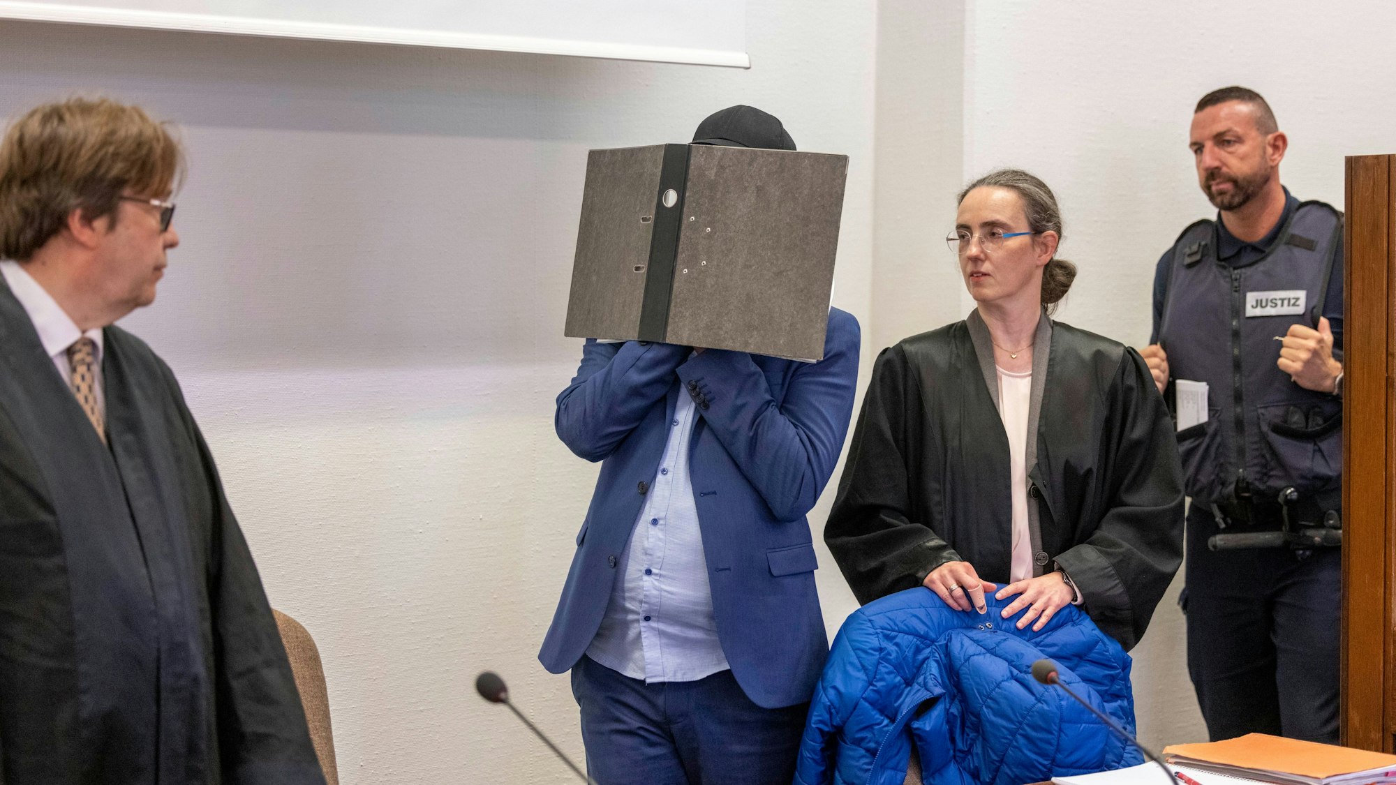 Der Angeklagte kommt vor der Verkündung des Urteils im Prozess wegen sexuellen Missbrauchs an Kindern in den Gerichtssaal des Kölner Landgerichts, während er sein Gesicht hinter einem Aktenordner verbirgt. Seine beiden Rechtsanwälte Jürgen Schüttler (links) und Sybille Weber (rechts) erwarten den Angeklagten im Gerichtssaal.