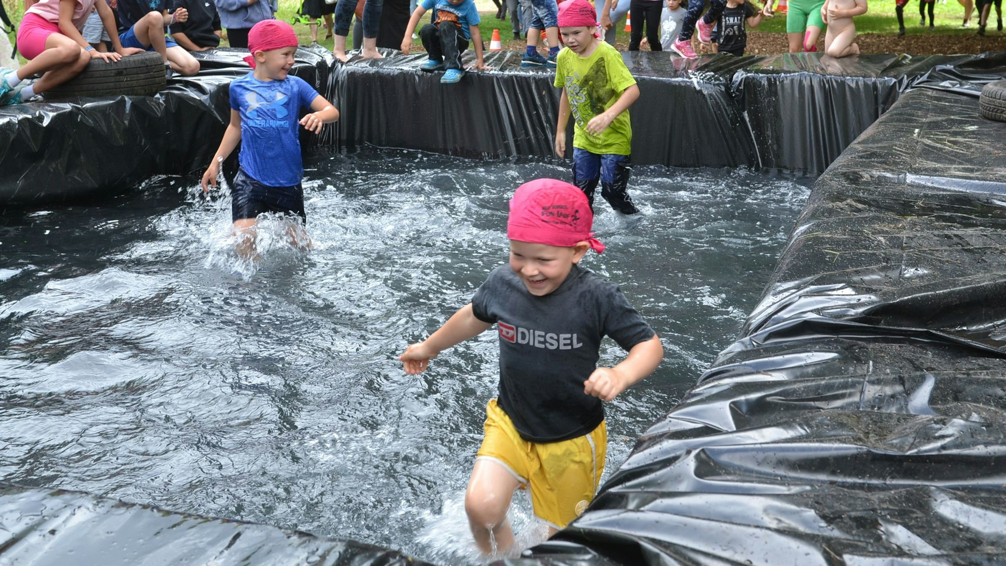 Drei Kinder laufen durch ein Wasserbecken. Das Wasser reicht ihnen bis zu den Knien. Sie tragen pinke Tücher um den Kopf und lachen.