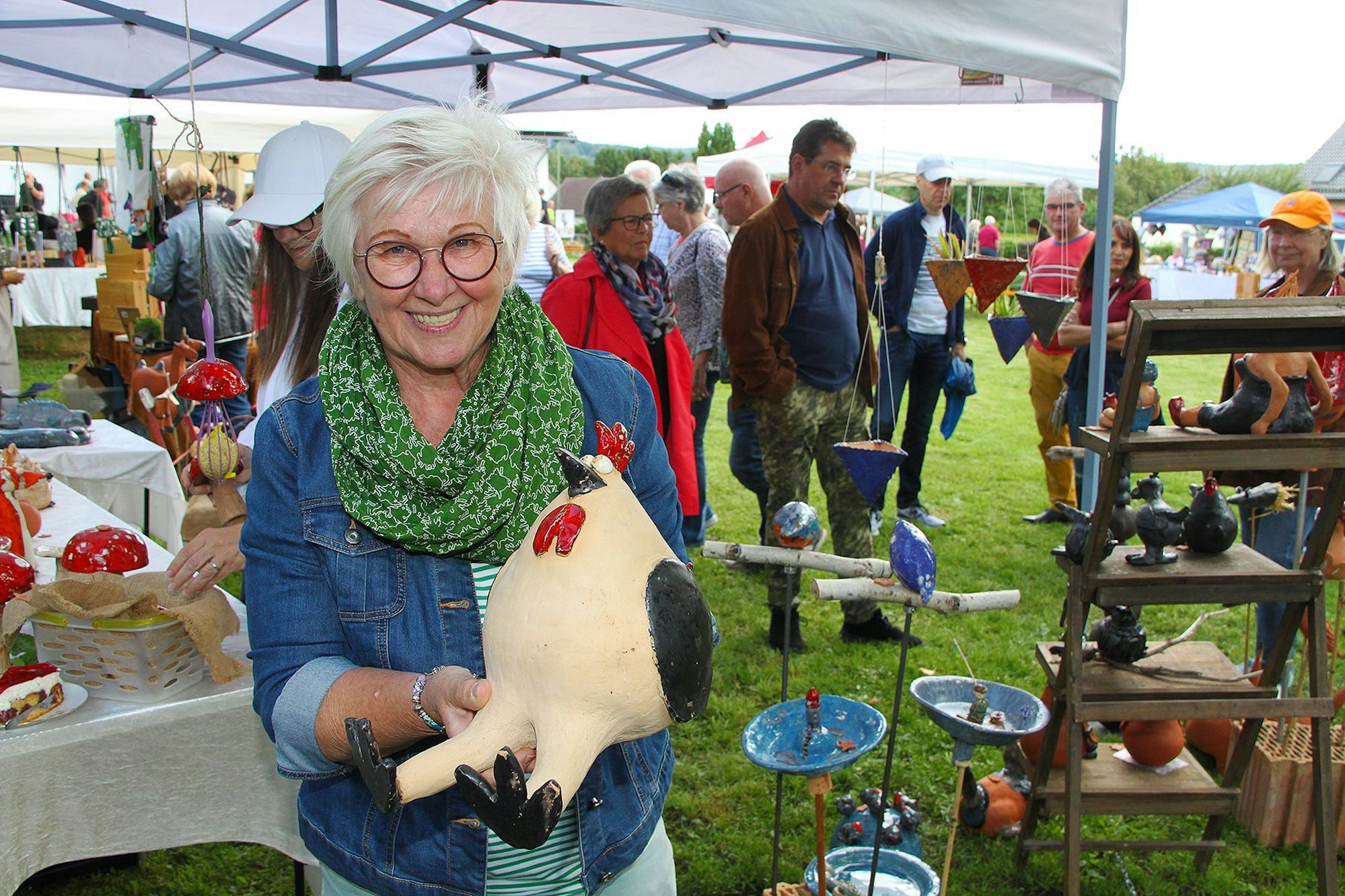 Eine Frau mit Brille und grauen Haaren hält ein dickes Huhn aus Keramik in die Kamera. Im Hintergrund sind Stände und Marktbesucher zu sehen.