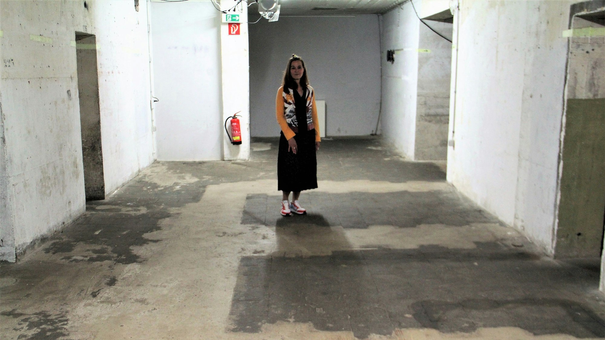Eine Frau mit langen dunklen Haaren steht in einem nackten kellerartigen Raum.