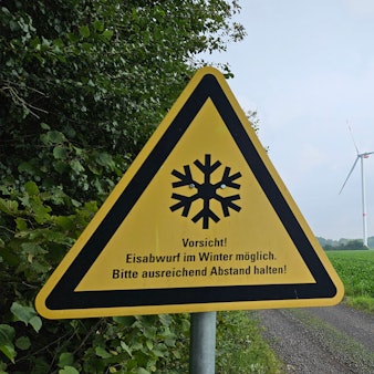 Ein Warnschild steht am Waldsaum neben einem Acker, im Hintergrund erheben sich drei Windräder.