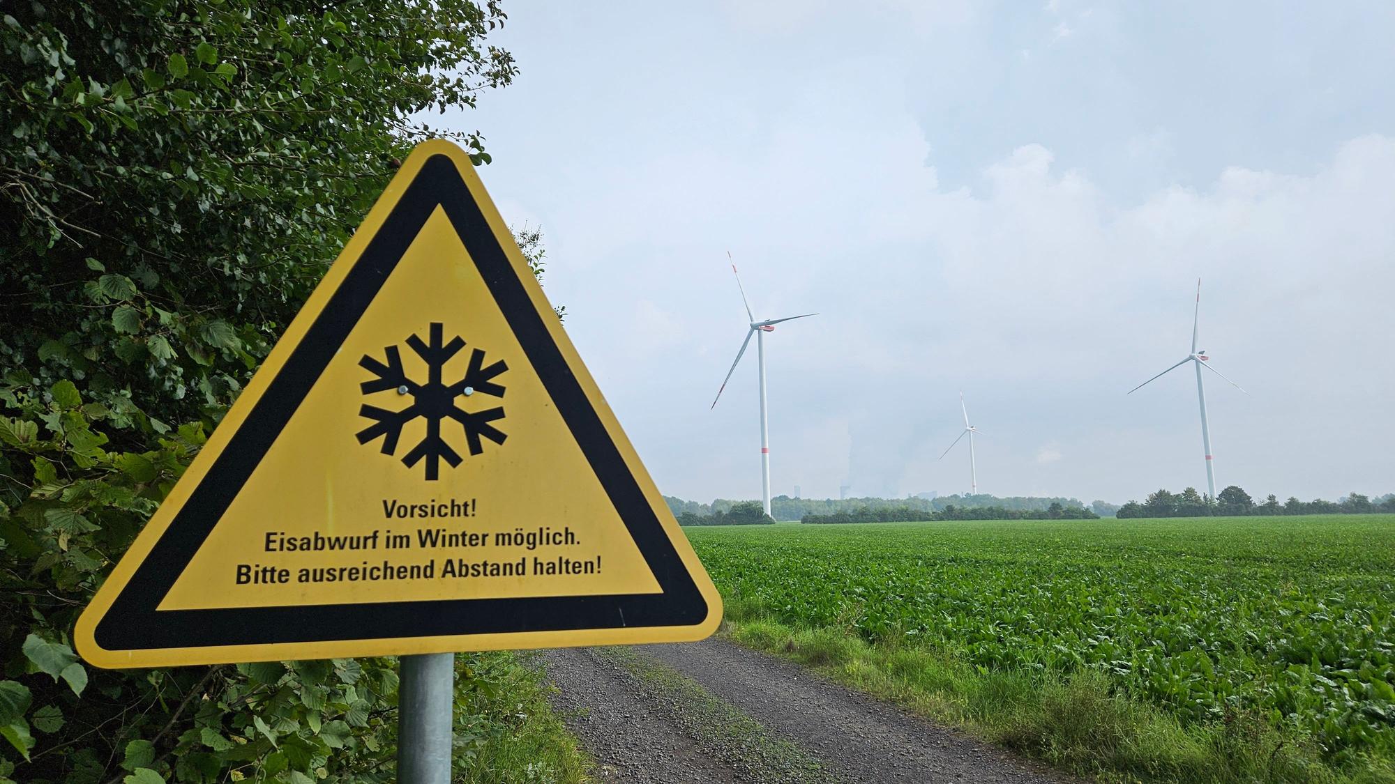 Ein Warnschild steht am Waldsaum neben einem Acker, im Hintergrund erheben sich drei Windräder.