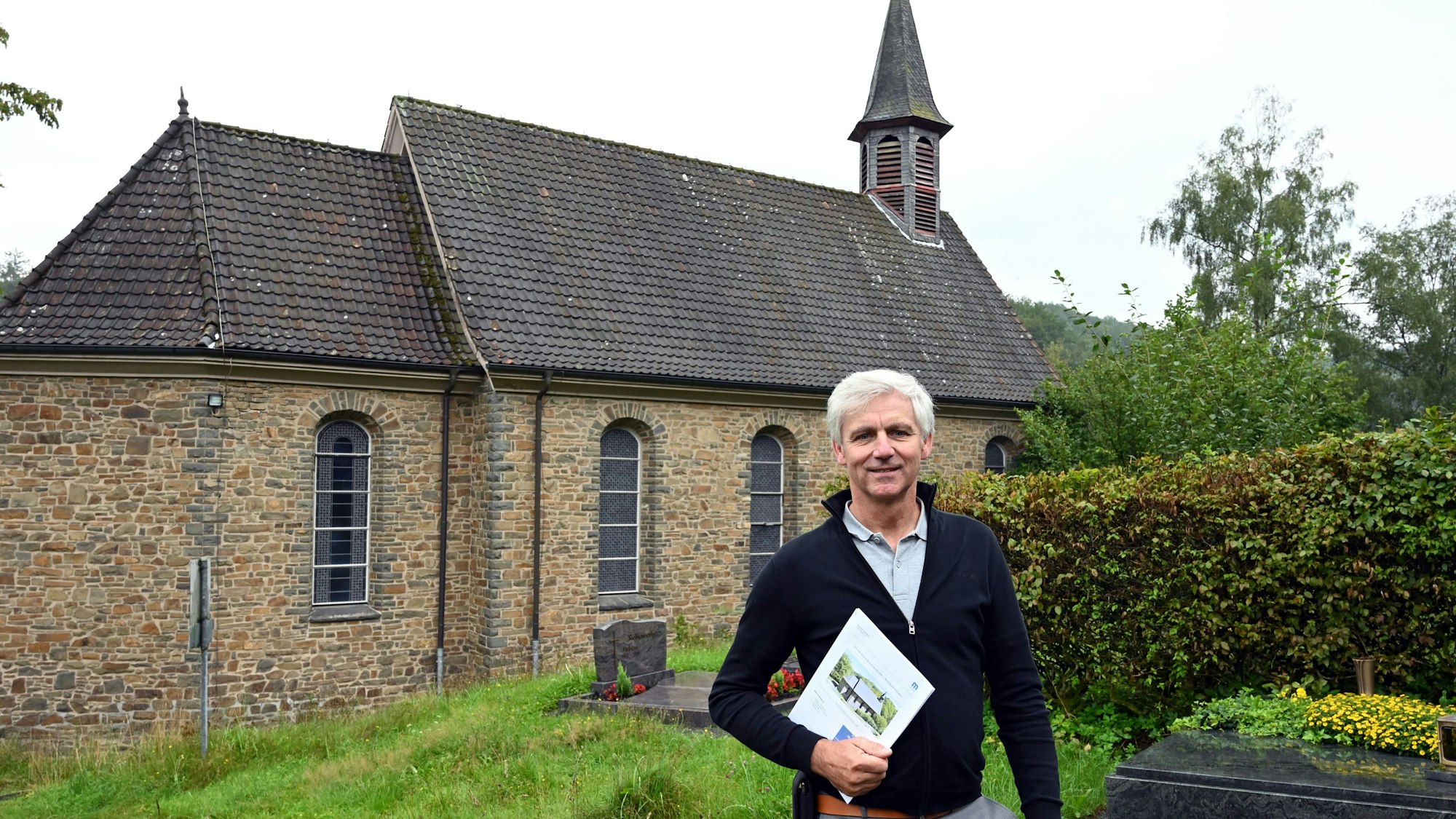 Der Morsbacher Makler Christoph Langer hat den Auftrag erhalten, die katholische Christ-König-Kirche in der Ortschaft Ellingen zu verkaufen. Hier steht er auf dem Friedhof der Kirche, der vorerst erhalten bleiben soll.