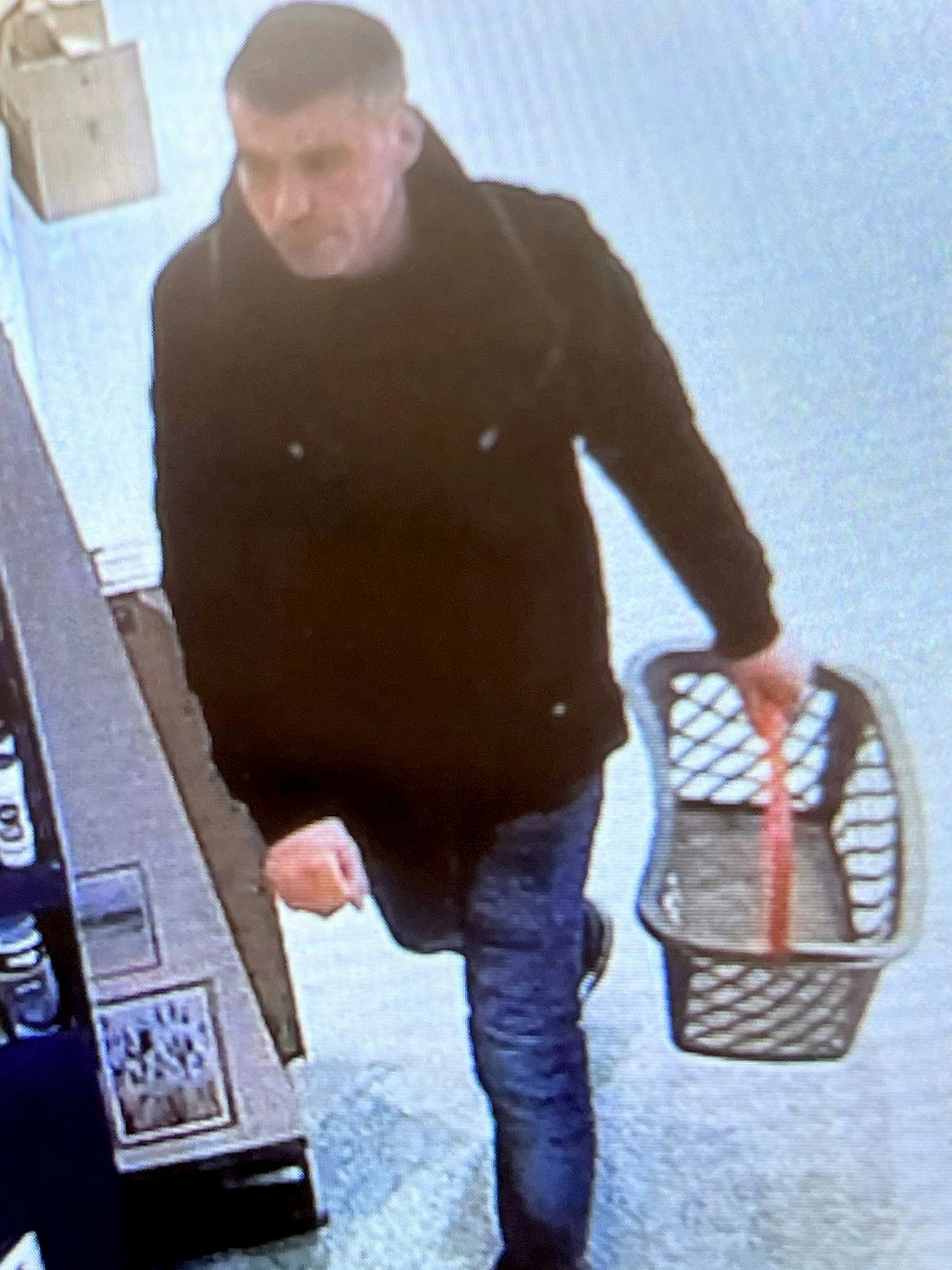 Ein Mann mit kurzen dunklen Haaren trägt einen leeren Einkaufskorb. Er ist mit einer schwarzen Jacke, blauen Jeans und schwarzen Turnschuhen bekleidet.