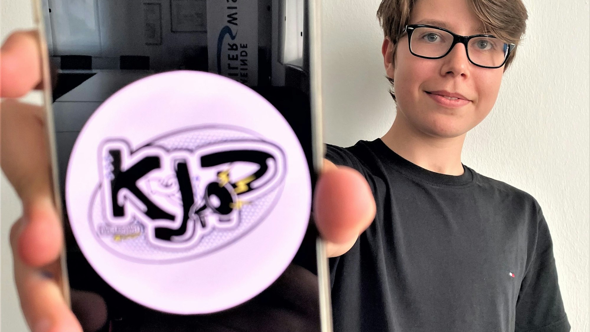 Simon Krämer, Vorsitzender des Kinder- und Jugendparlaments, hält sein Smartphone in die Kamera. Darauf zu sehen: das Logo des Kinder- und Jugendparlaments (KJP).