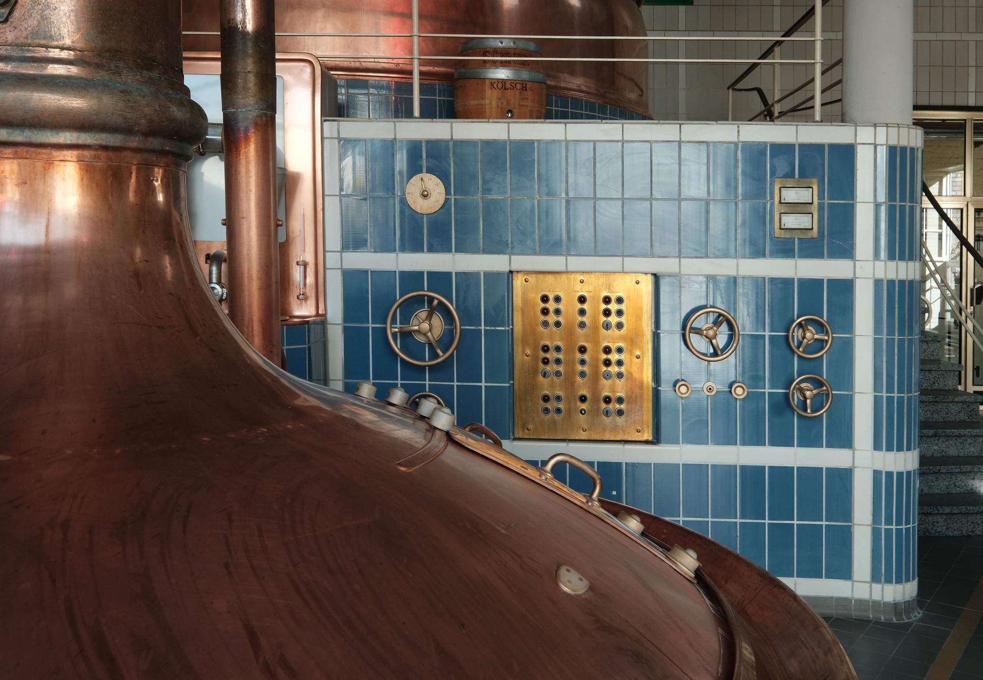 Zu sehen ist eine bronzene Sudpfanne und dahinter ein blau geflieste Wand mit allerlei rundem Hebeln, die aussehen wie kleine Lenkräder.