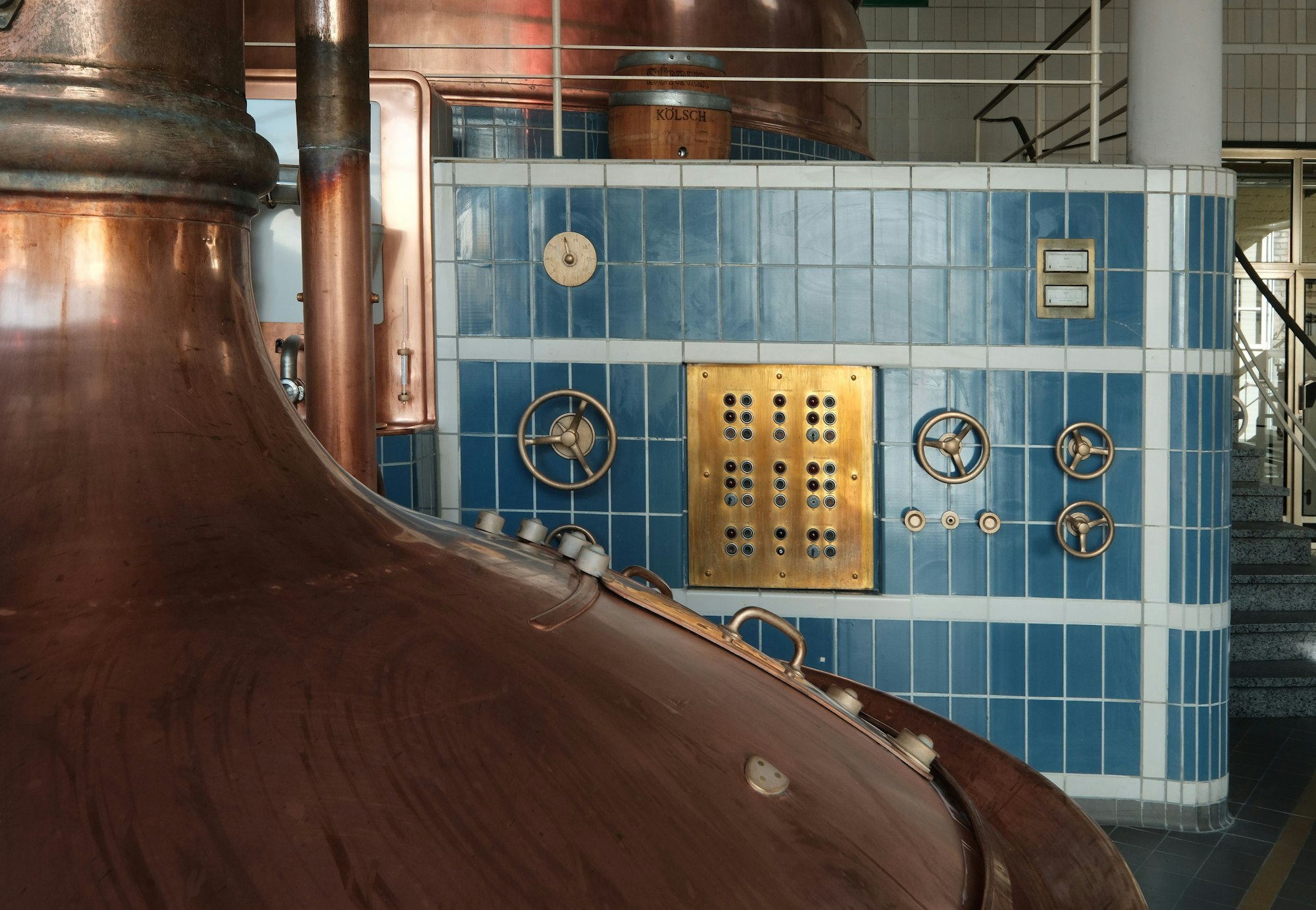 Zu sehen ist eine bronzene Sudpfanne und dahinter ein blau geflieste Wand mit allerlei rundem Hebeln, die aussehen wie kleine Lenkräder.