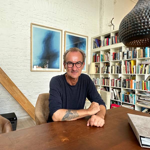 Ein Mann mit hoher Stirn, schwarzer Brille und im blauen Pullover sitzt an einem Tisch, im Hintergrund sind Bilder und ein Bücherregal zu sehen.