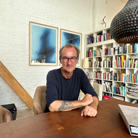Ein Mann mit hoher Stirn, schwarzer Brille und im blauen Pullover sitzt an einem Tisch, im Hintergrund sind Bilder und ein Bücherregal zu sehen.
