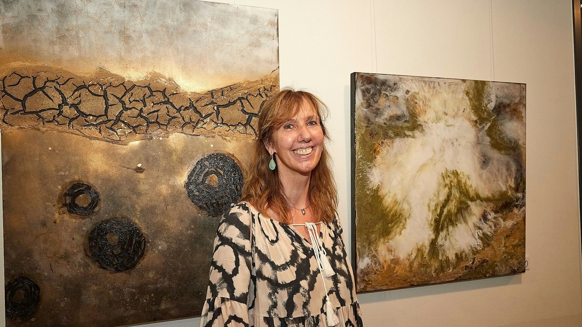 Im Rathaus in Mechernich steht Ela Rübenach vor zwei ihrer Gemälde, die mit Kunstharz, Acryl und anderen Materialien geschaffen wurden. Die Bilder sehen aus wie Baumrinde, Verbranntes und Ausgelaufenes.