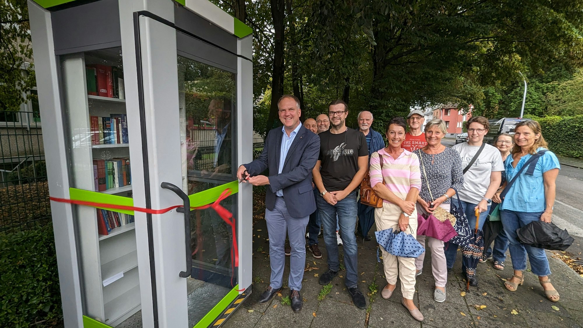 Auf dem Foto ist zu sehen, wie Hürths Bürgermeister Dirk Breuer das Band durchschneidet und den neuen Bücherschrank vor der Brüder-Grimm-Schule freigibt.