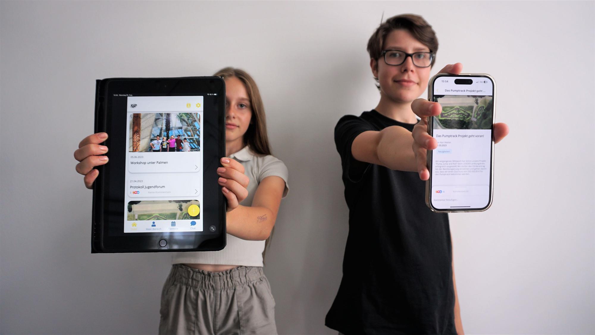 Sophie Schmidt und Simon Krämer halten mit ausgestreckten Armen ein Tablet und ein Smartphone in die Kamera