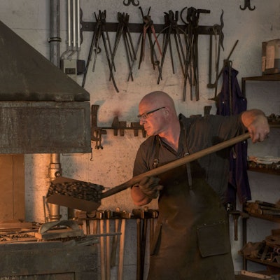 Der Schmied schaufelt gerade heiße Kohlen in die Schmiede, in der ein Feuer brennt.