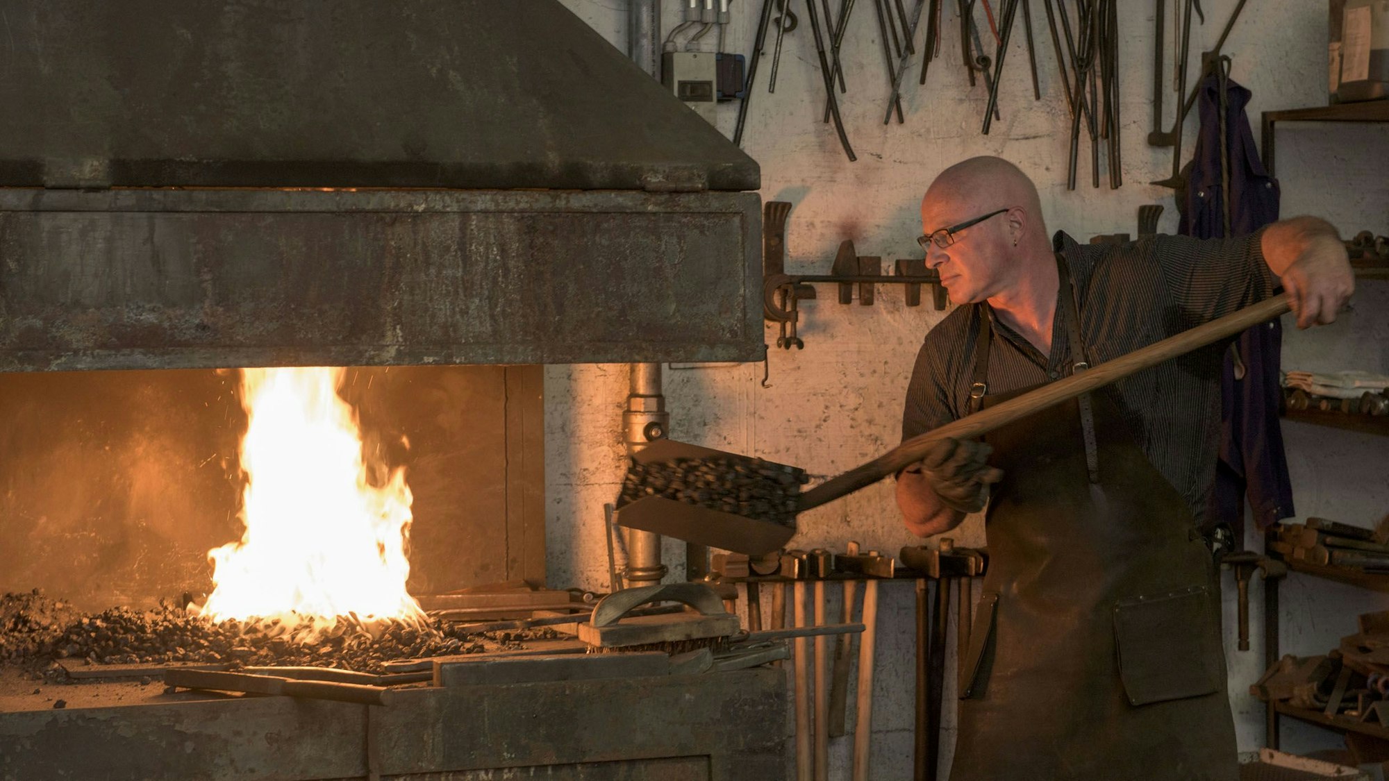 Der Schmied schaufelt gerade heiße Kohlen in die Schmiede, in der ein Feuer brennt.