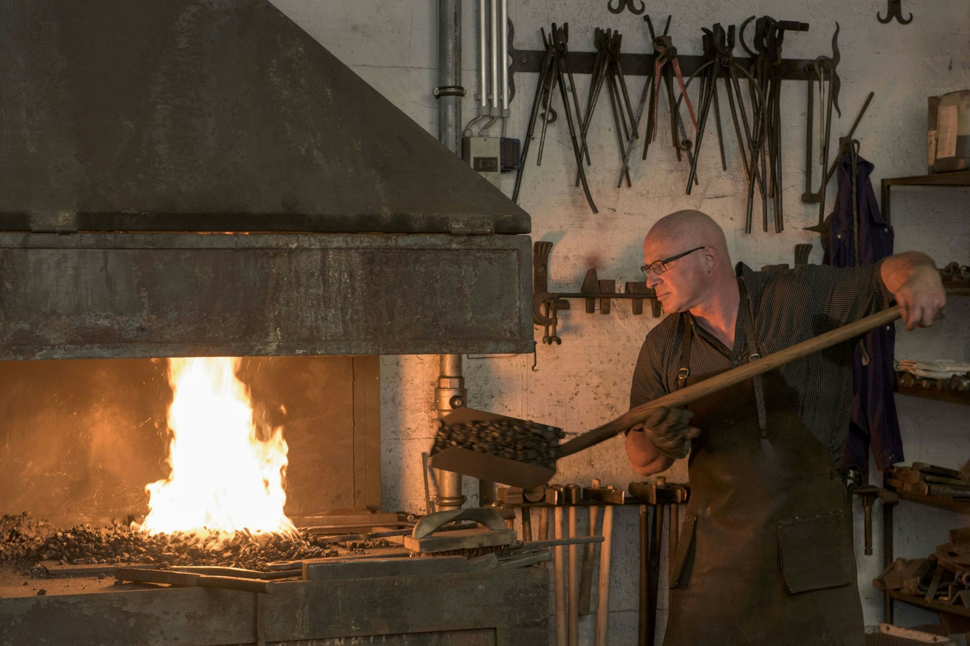 Der Schmied schaufelt gerade heiße Kohlen in die Schmiede, in der ein Feuer brennt.