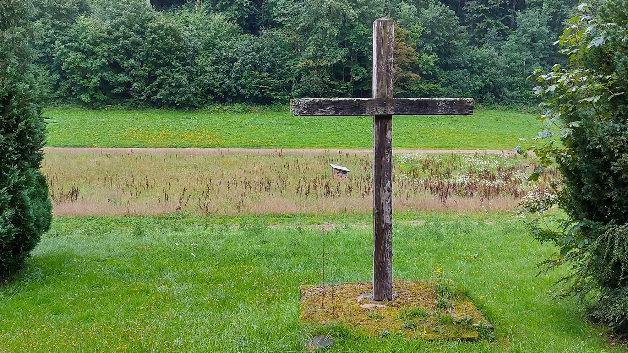 Ein Holzkreuz steht auf einer Rasenfläche, wo sich einst ein Friedhof befunden hat.