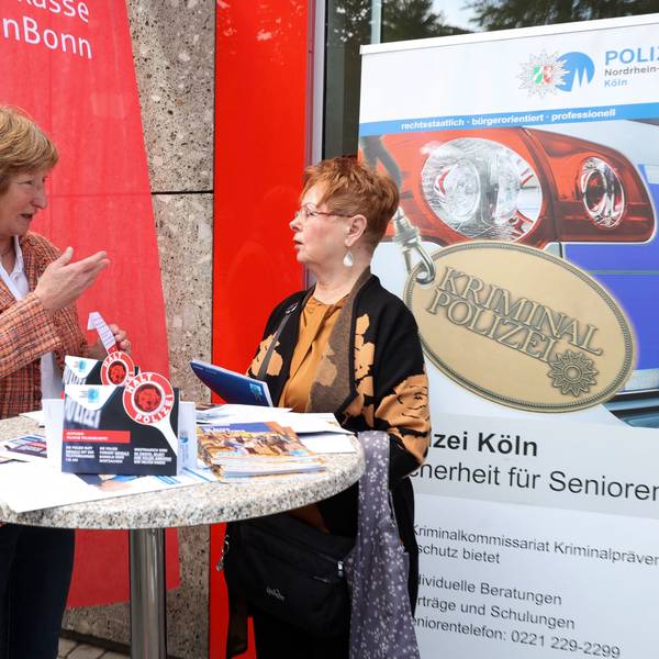 Gudrun Krämer, pensionierte Kriminalpolizistin, und Sparkassen-Kundin Renate Heinrichs stehen an einem Stehtisch vor der Sparkassen-Filiale in Köln-Porz und unterhalten sich.