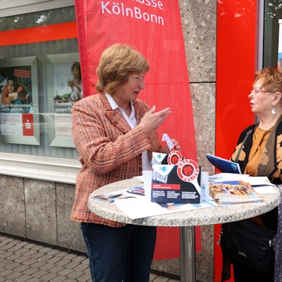 Gudrun Krämer, pensionierte Kriminalpolizistin, und Sparkassen-Kundin Renate Heinrichs stehen an einem Stehtisch vor der Sparkassen-Filiale in Köln-Porz und unterhalten sich.
