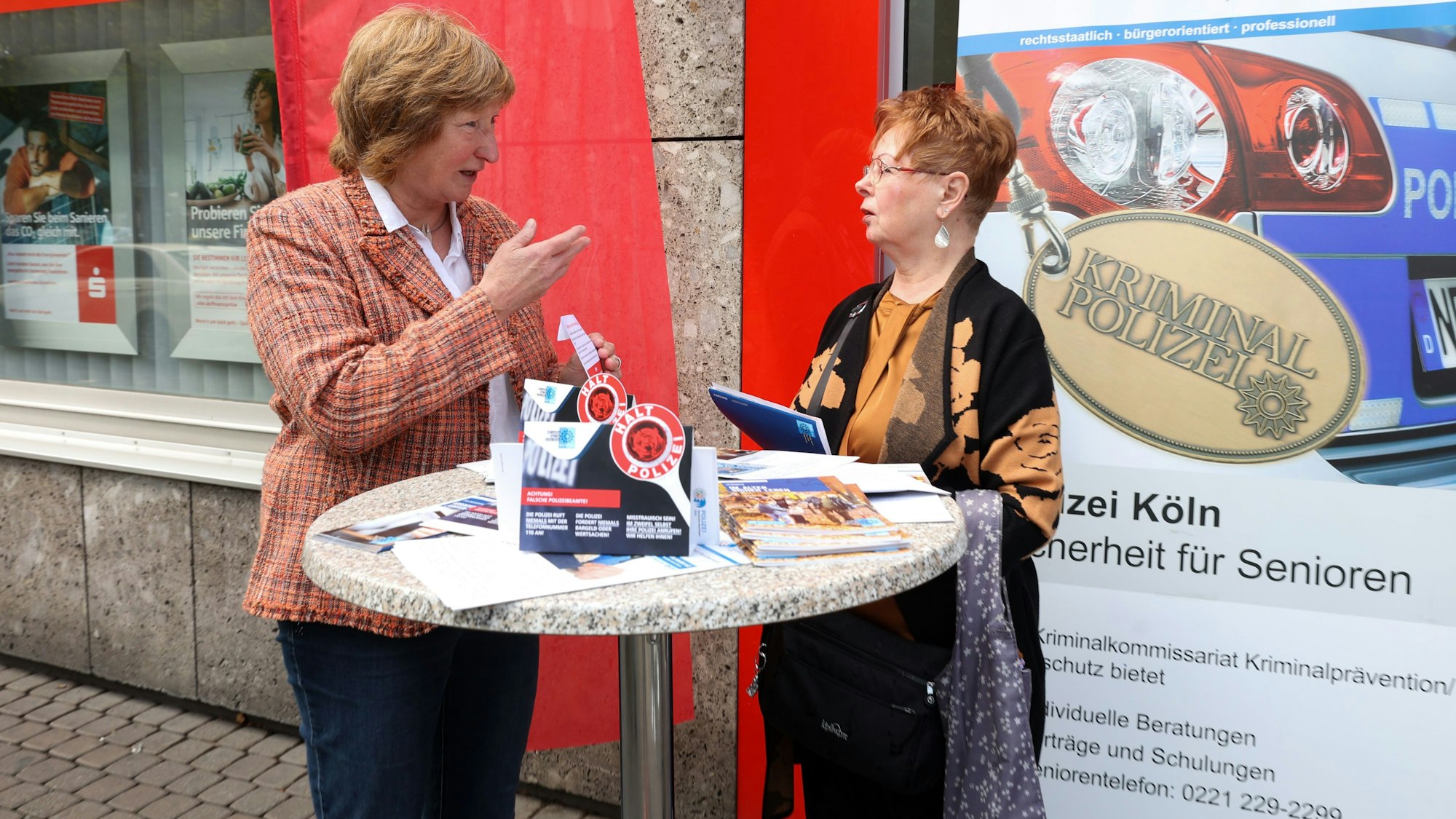 Gudrun Krämer, pensionierte Kriminalpolizistin, und Sparkassen-Kundin Renate Heinrichs stehen an einem Stehtisch vor der Sparkassen-Filiale in Köln-Porz und unterhalten sich.