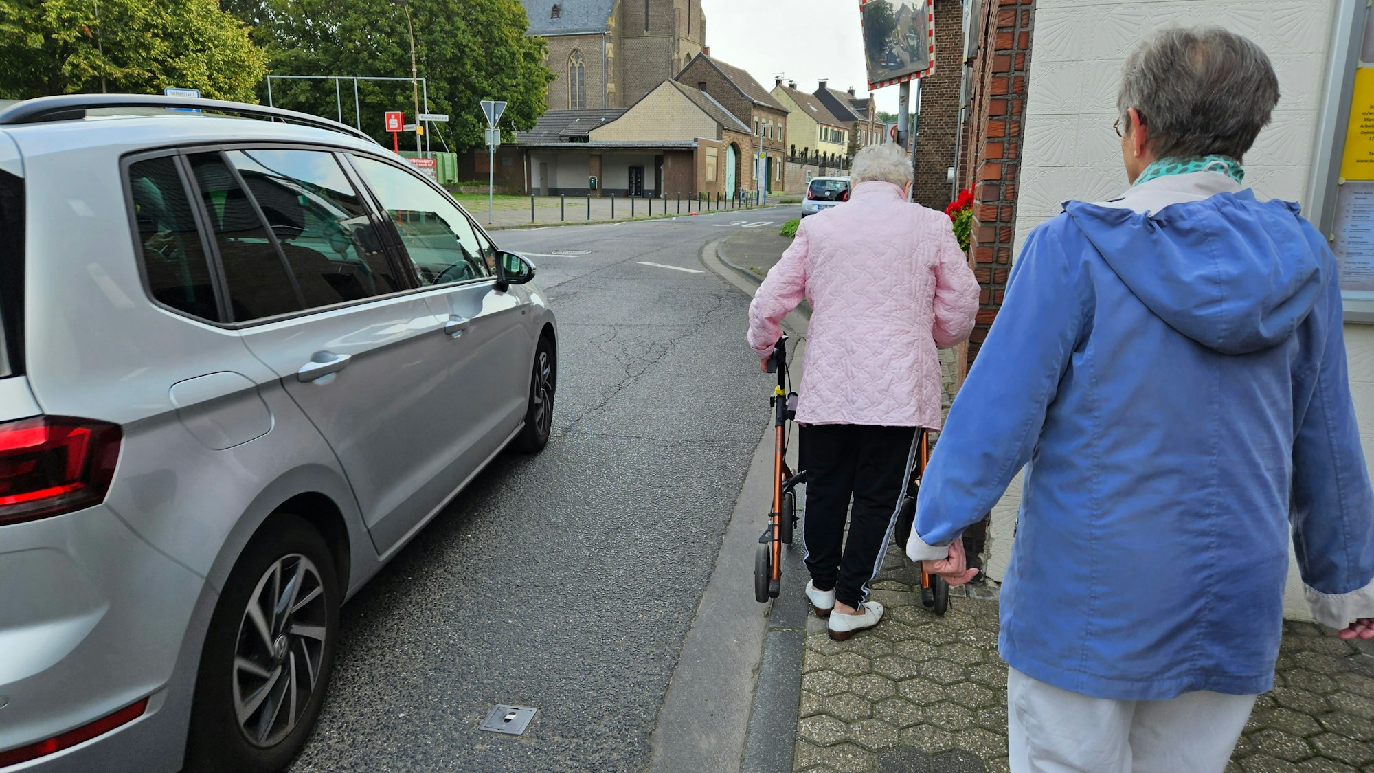 Zwei ältere Frauen, eine an einer rollbaren Gehhilfe, gehen auf einem schmalen Gehweg, ein Auto fährt an ihnen vorbei.