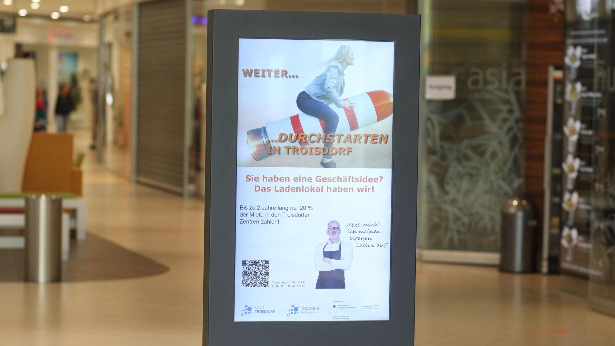 In einer Einkaufspassage steht ein beleuchtetes Plakat, auf dem eine rFau auf einer schräg nach oben abhebenden Rakete sitzt.