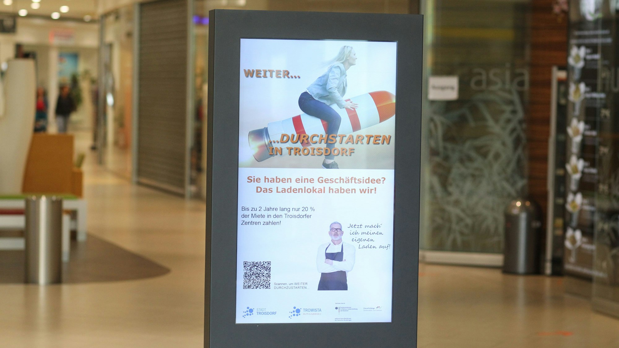 In einer Einkaufspassage steht ein beleuchtetes Plakat, auf dem eine rFau auf einer schräg nach oben abhebenden Rakete sitzt.