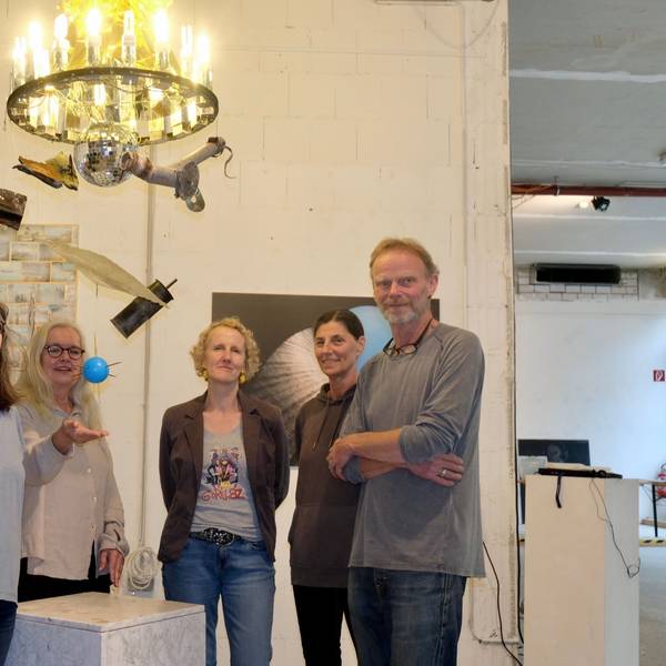 Vier Frauen und ein Mann stehen unter einem Kronleuchter, unter dem eine Diskokugel hängt, noch weiter unten baumeln eine blaue Kugel und verschiedene Objekte.