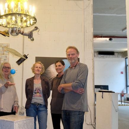 Vier Frauen und ein Mann stehen unter einem Kronleuchter, unter dem eine Diskokugel hängt, noch weiter unten baumeln eine blaue Kugel und verschiedene Objekte.