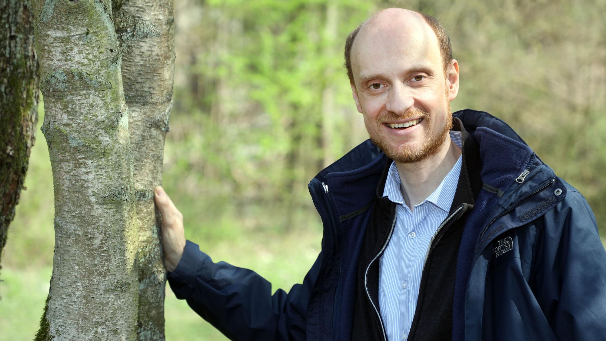 Der Biologe Dr. Bernd Freymann steht neben einem Baum und legt eine Hand auf den Stamm.