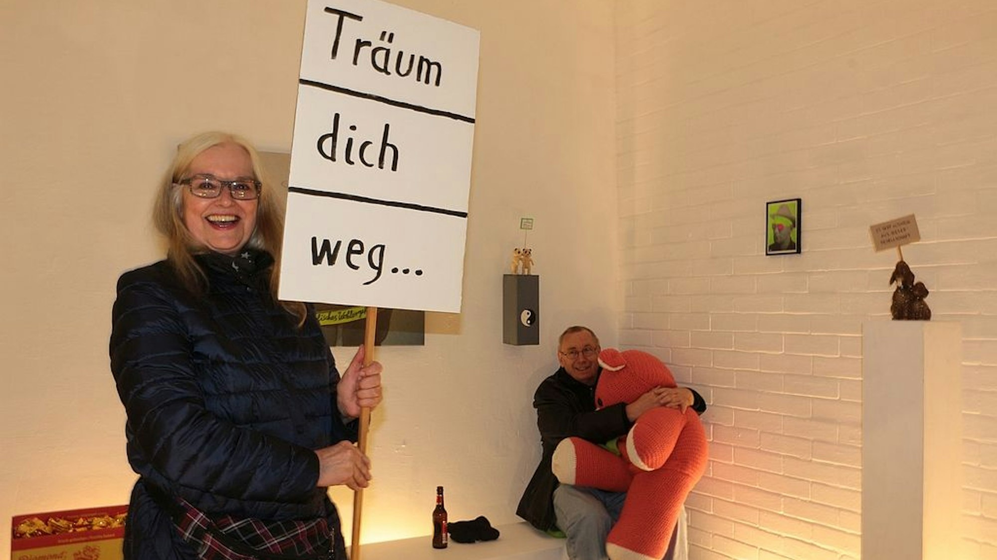 Eine Frau hält ein Schild mit der Aufforderung Träum dich weg. Im Hintergrund ist ein Mann auf einer Bank zu sehen, mit einem riesigen Teddybär auf dem Schoß.